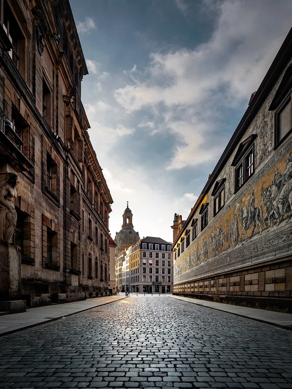 Cobblestone street in Dresden leading to Frauenkirche dome, flanked by old buildings and the Fürstenzug mural on the right.