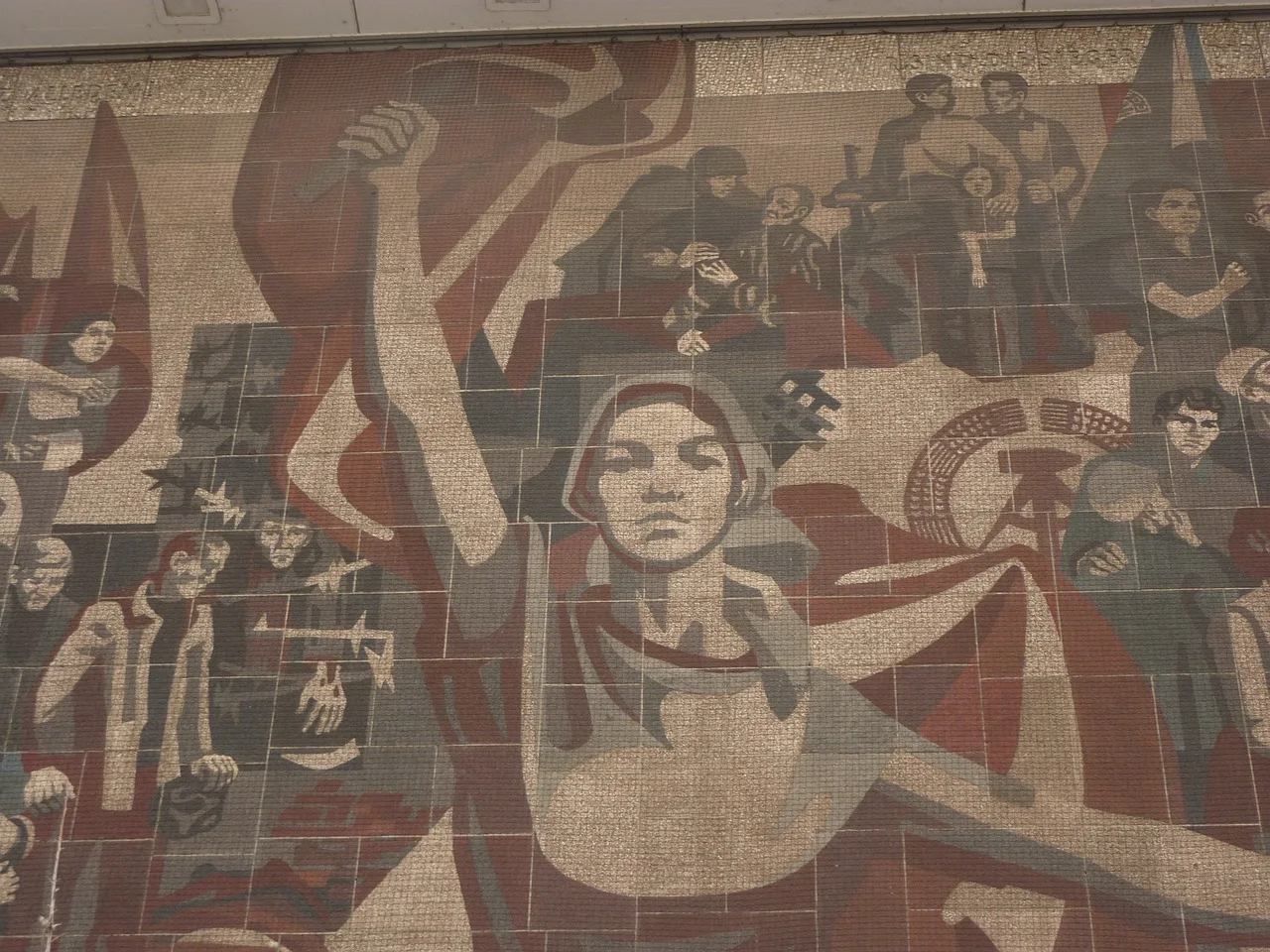 Gran mural de mosaico realista-socialista en tonos rojos, grises y beige, que representa trabajadores, banderas y una figu...