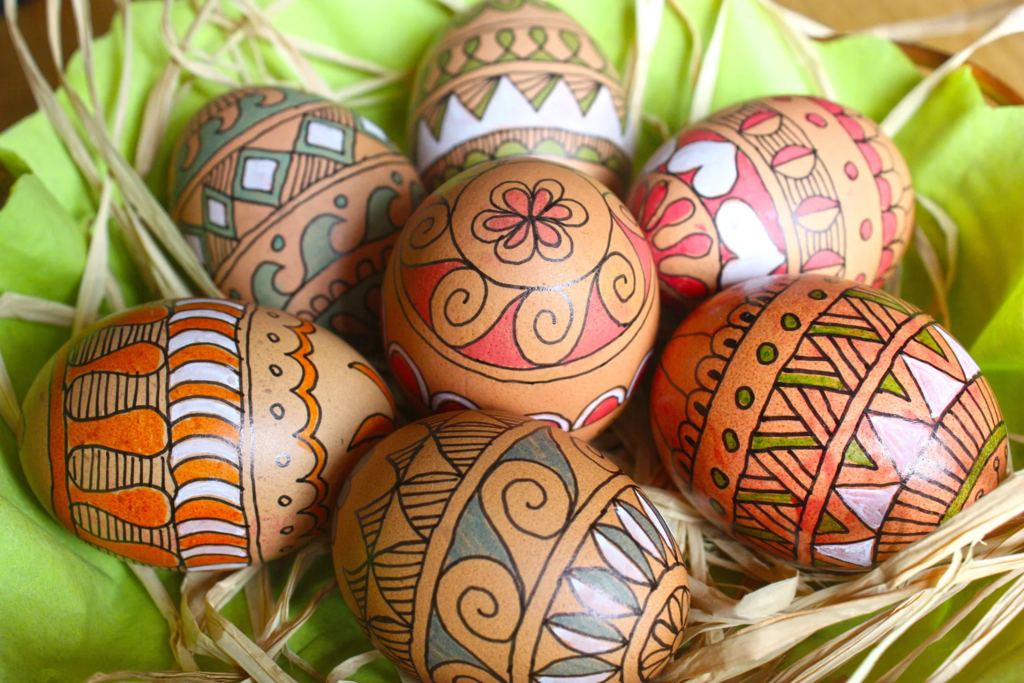 Seven brown Easter eggs with intricate red, green, orange, and white patterns, resting in a nest of straw and green fabric.