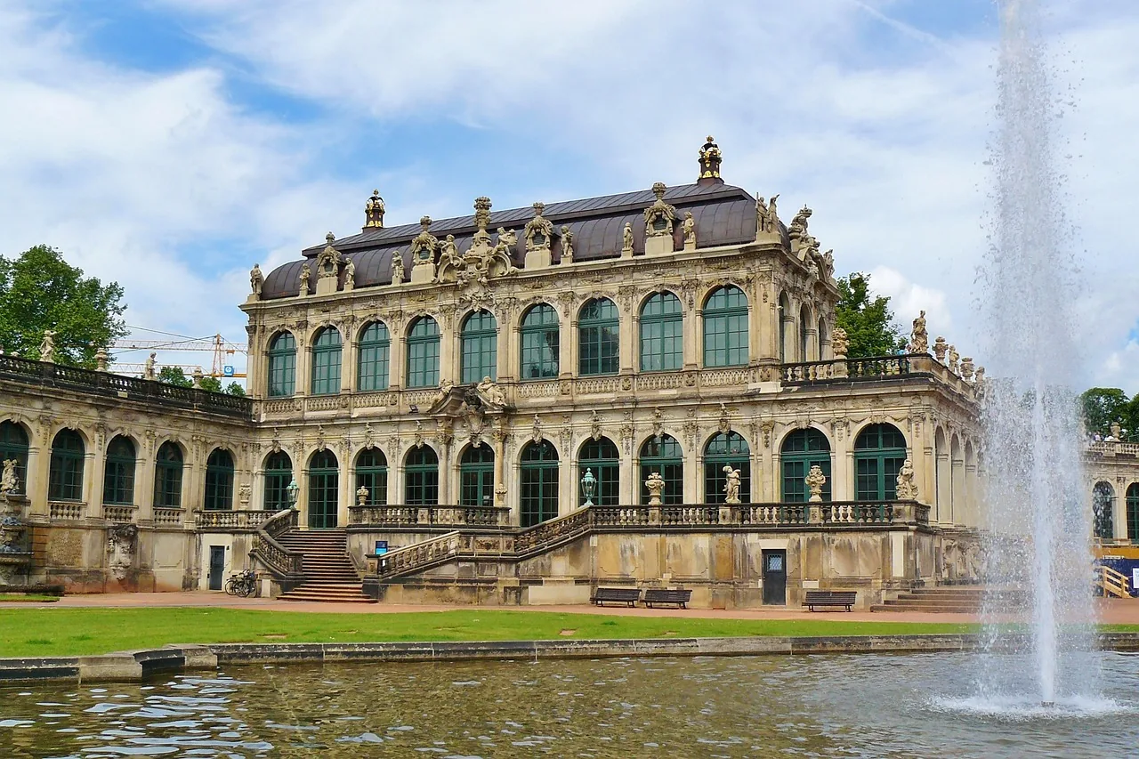 Barocke Palastfassade mit grün gerahmten Fenstern und opulenten Details, gesehen über einen Teich mit großer Fontäne.