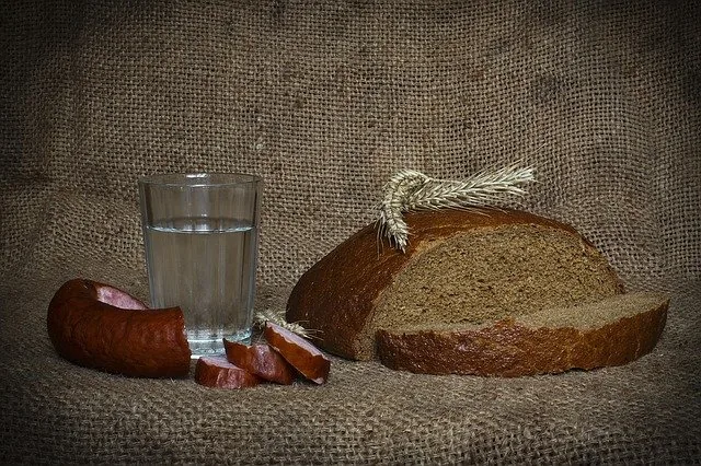 Vaso de líquido transparente, rebanadas de pan de centeno oscuro con espigas de trigo y salchicha cortada sobre arpillera.