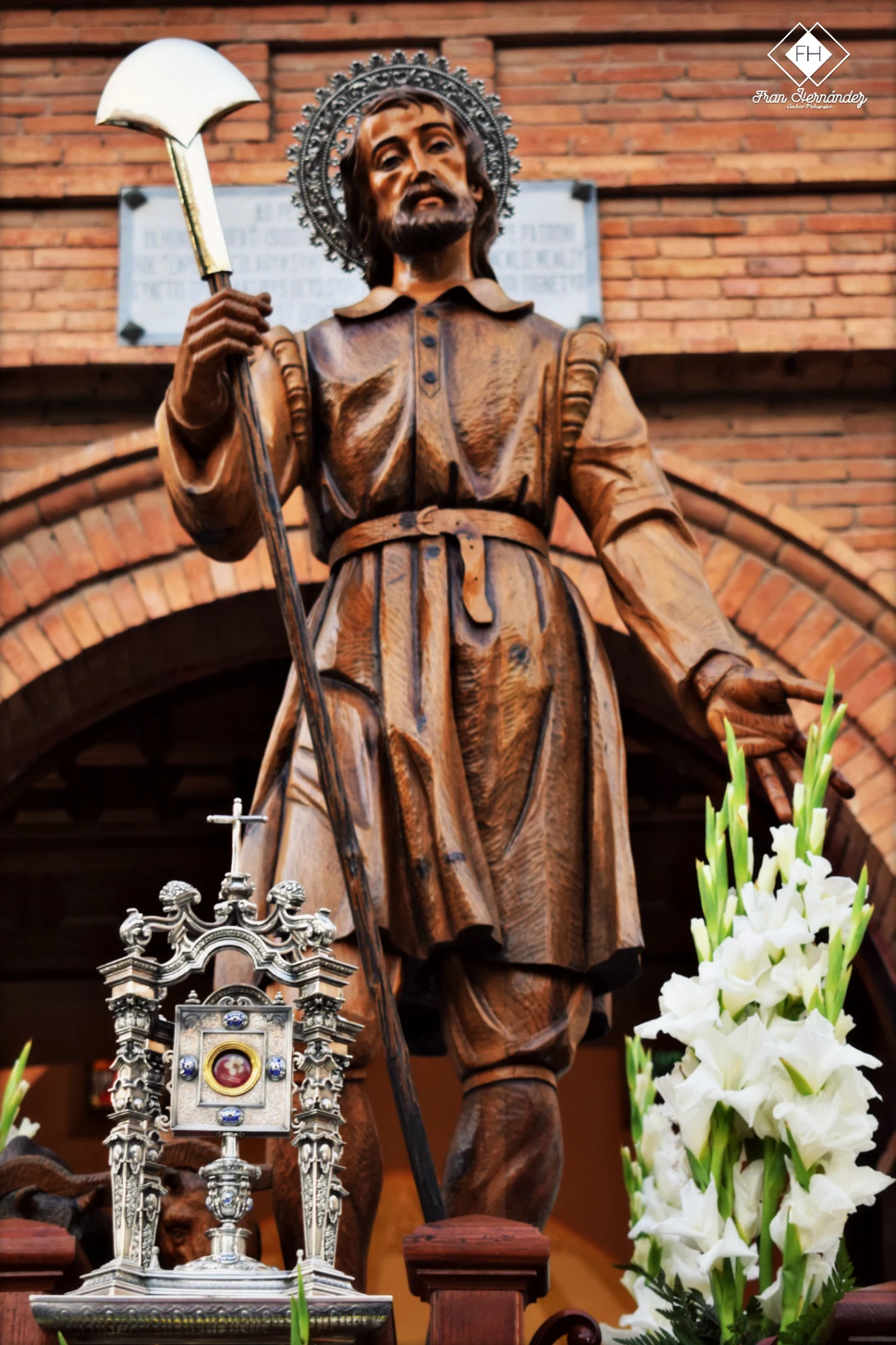A wooden statue of Saint Isidore the Laborer with a halo, holding a spade. In front is a reliquary and white flowers.
