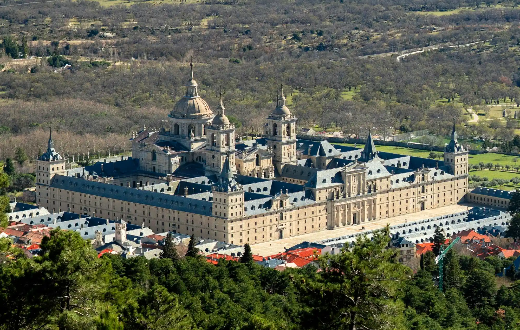 Aerial view of the San Lorenzo de El Escorial Royal Site, a large stone monastery complex with domes and spires, surrounded b