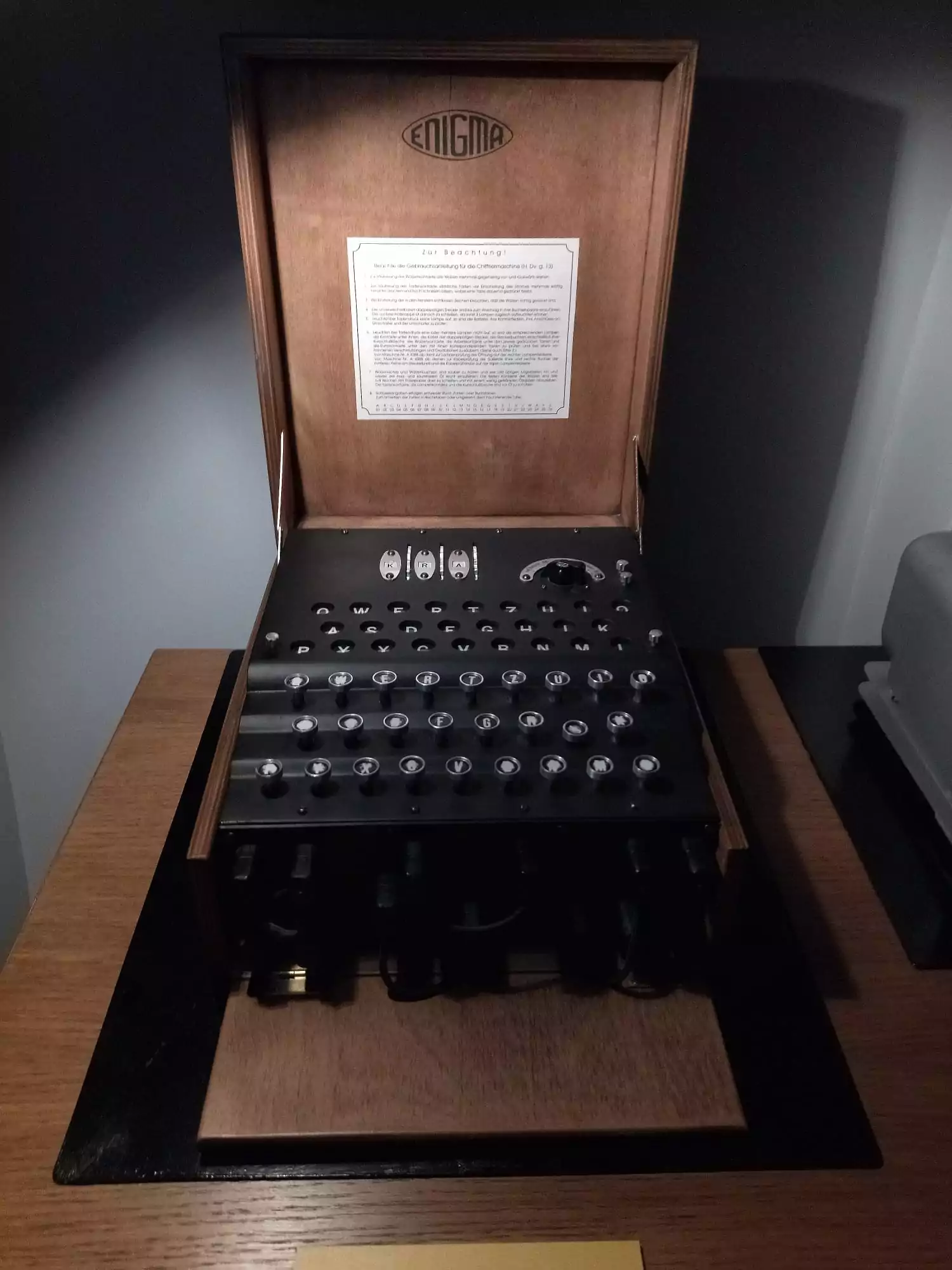 An open wooden case revealing a black Enigma machine with a keyboard and three letter displays. A document is on the inside l