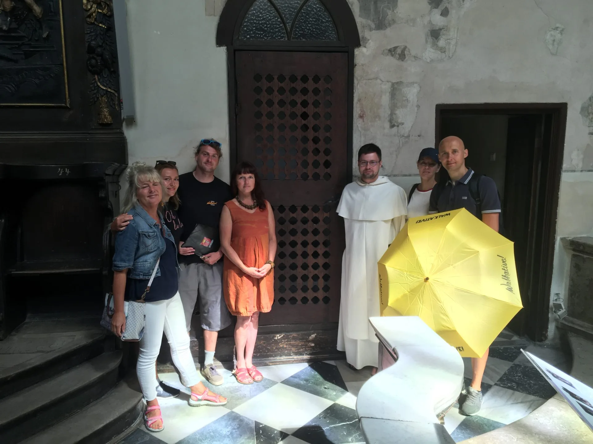 Siedem osób, w tym mężczyzna w habicie zakonnym, stoi we wnętrzu kościoła z ciemnymi drzwiami i szachownicową posadzką. Widoc
