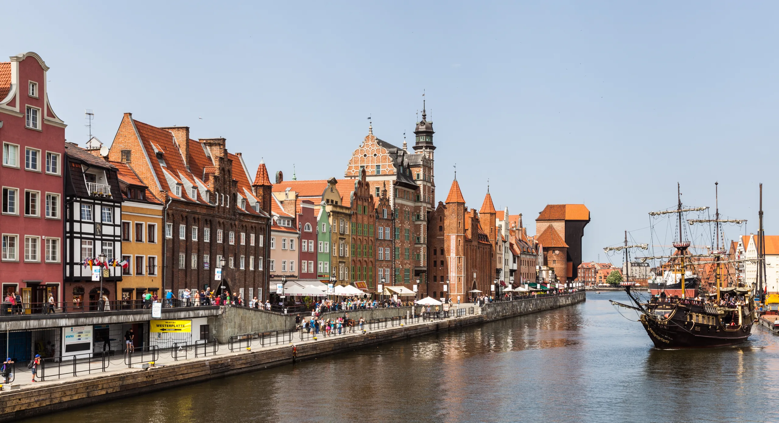 Historische Ufergebäude entlang des Flusses Mottlau in Danzig, mit Passanten und einem großen Segelschiff. Das Krantor ist si