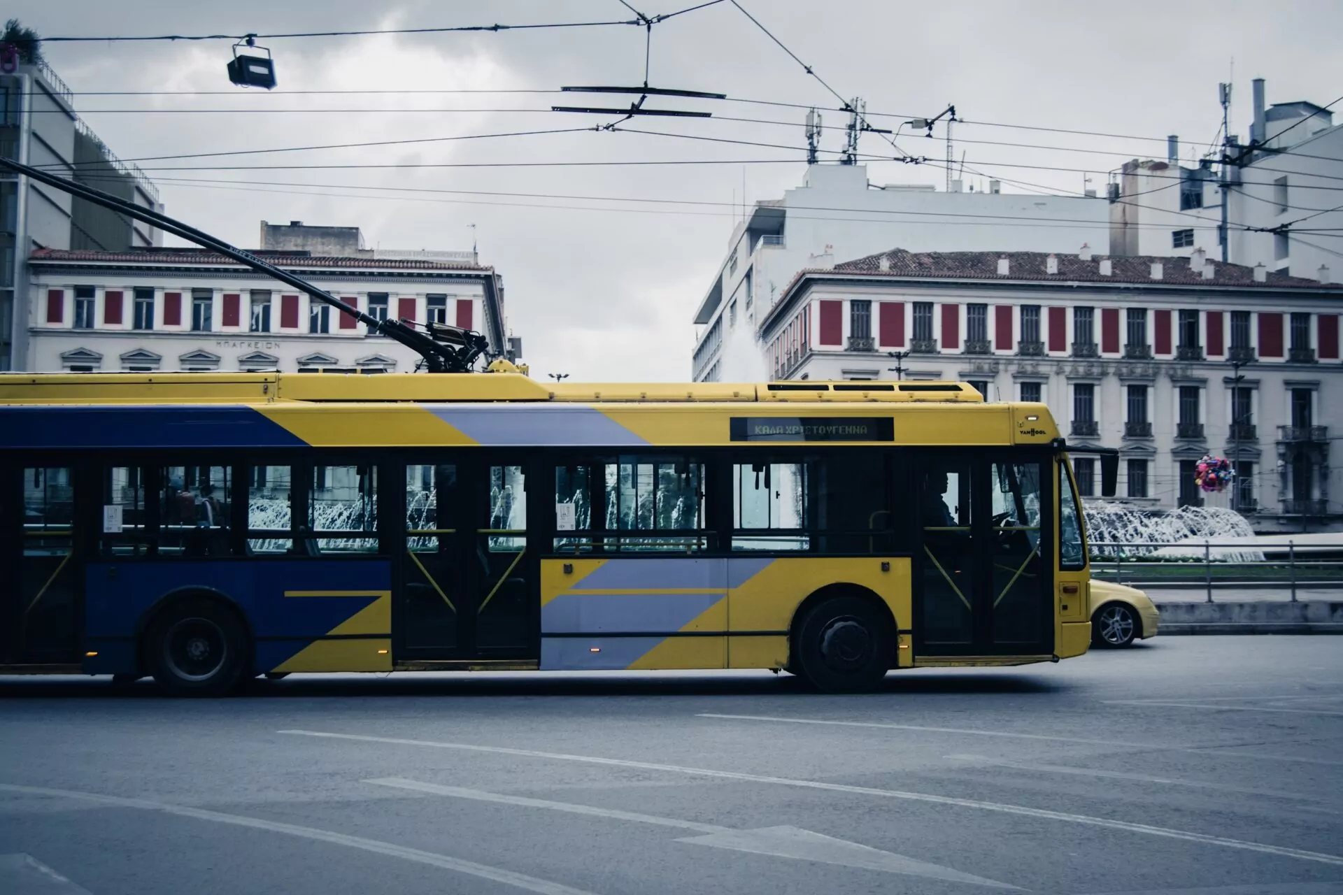 Yellow and blue electric trolleybus moving on a city street in Athens, with buildings and overhead lines against a cloudy sky