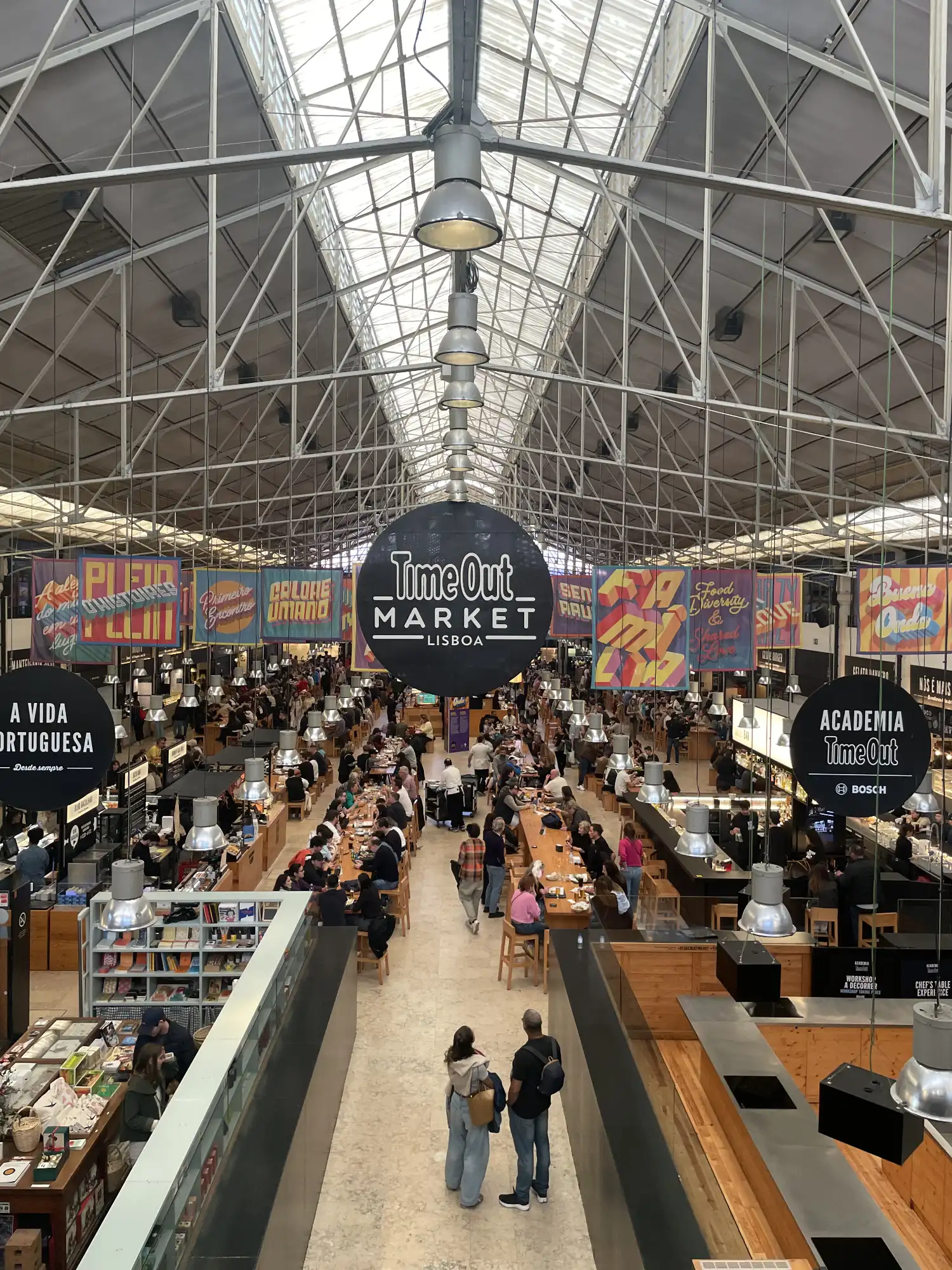 View from above of Time Out Market Lisboa, a bustling indoor food hall with many people dining at long tables and various foo
