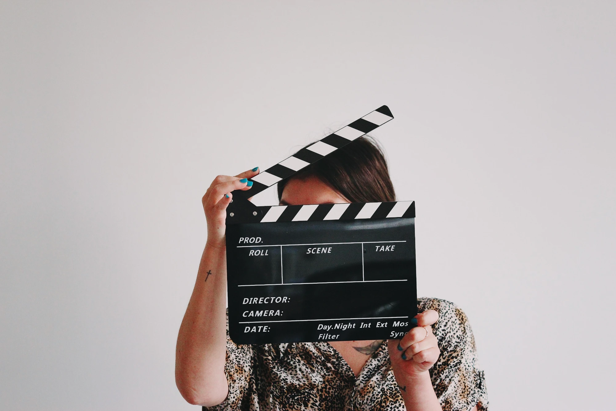 A woman holds a film clapperboard in front of her face. She has blue nail polish and a small cross tattoo on her arm.