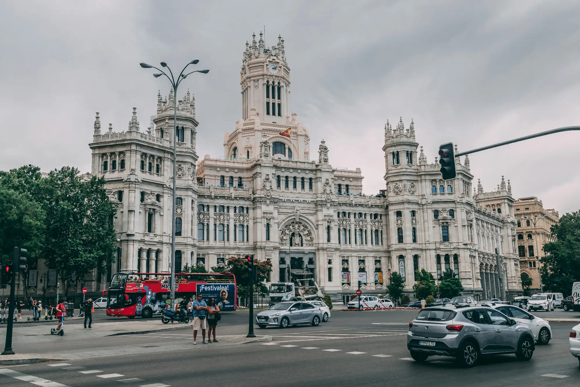 The grand white facade of the Palacio de Cibeles in Madrid, with a busy street scene of cars and a tour bus in front.