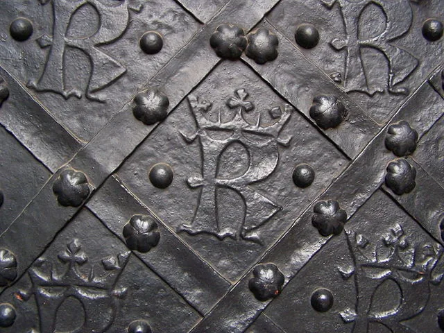 Nahaufnahme einer dunklen, verzierten Metalltür mit wiederholten gekrönten „R“-Monogrammen und Ziernieten in Rautenmuster.