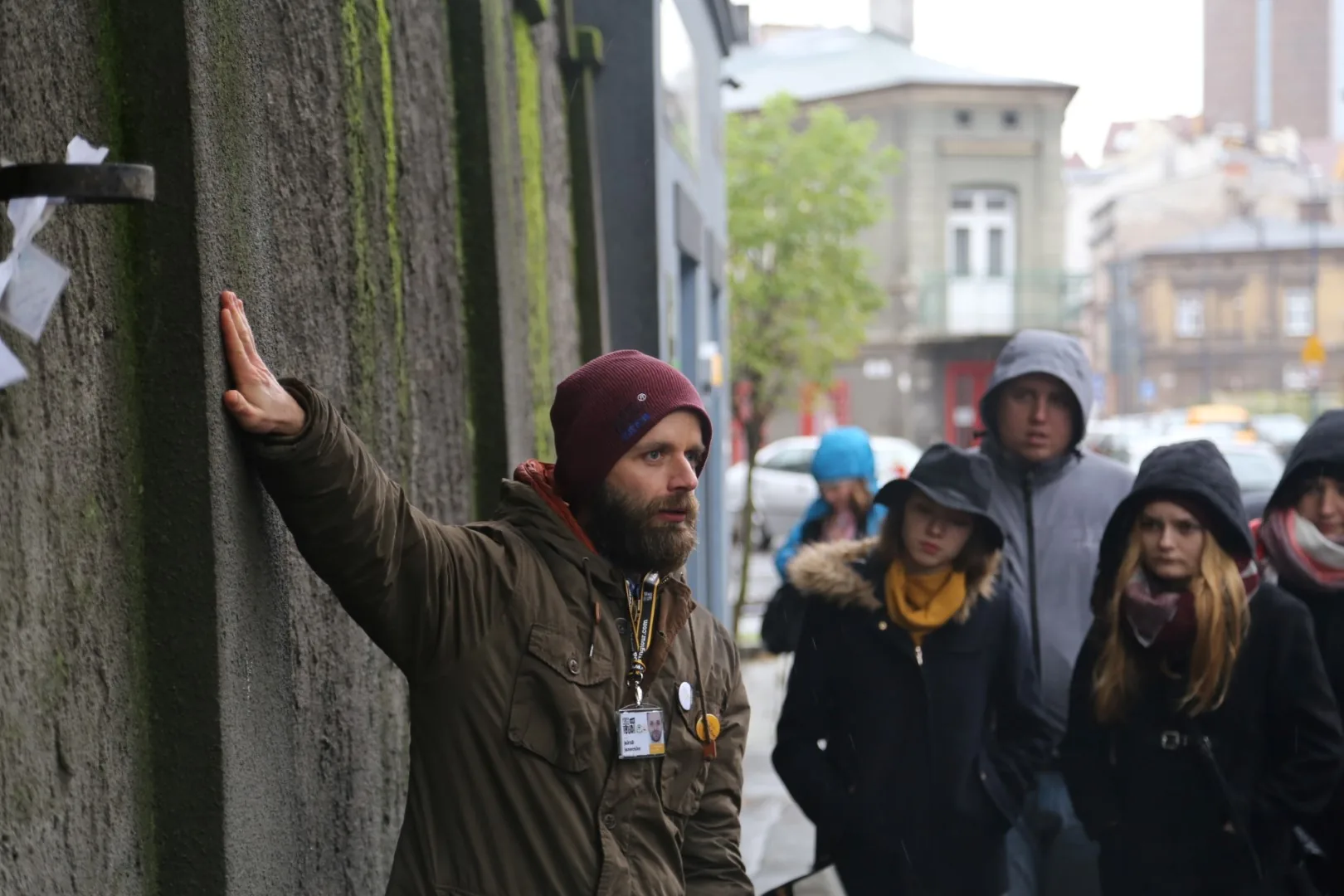 Tour guide with beard and beanie explaining to students by a mossy wall in Krakow.