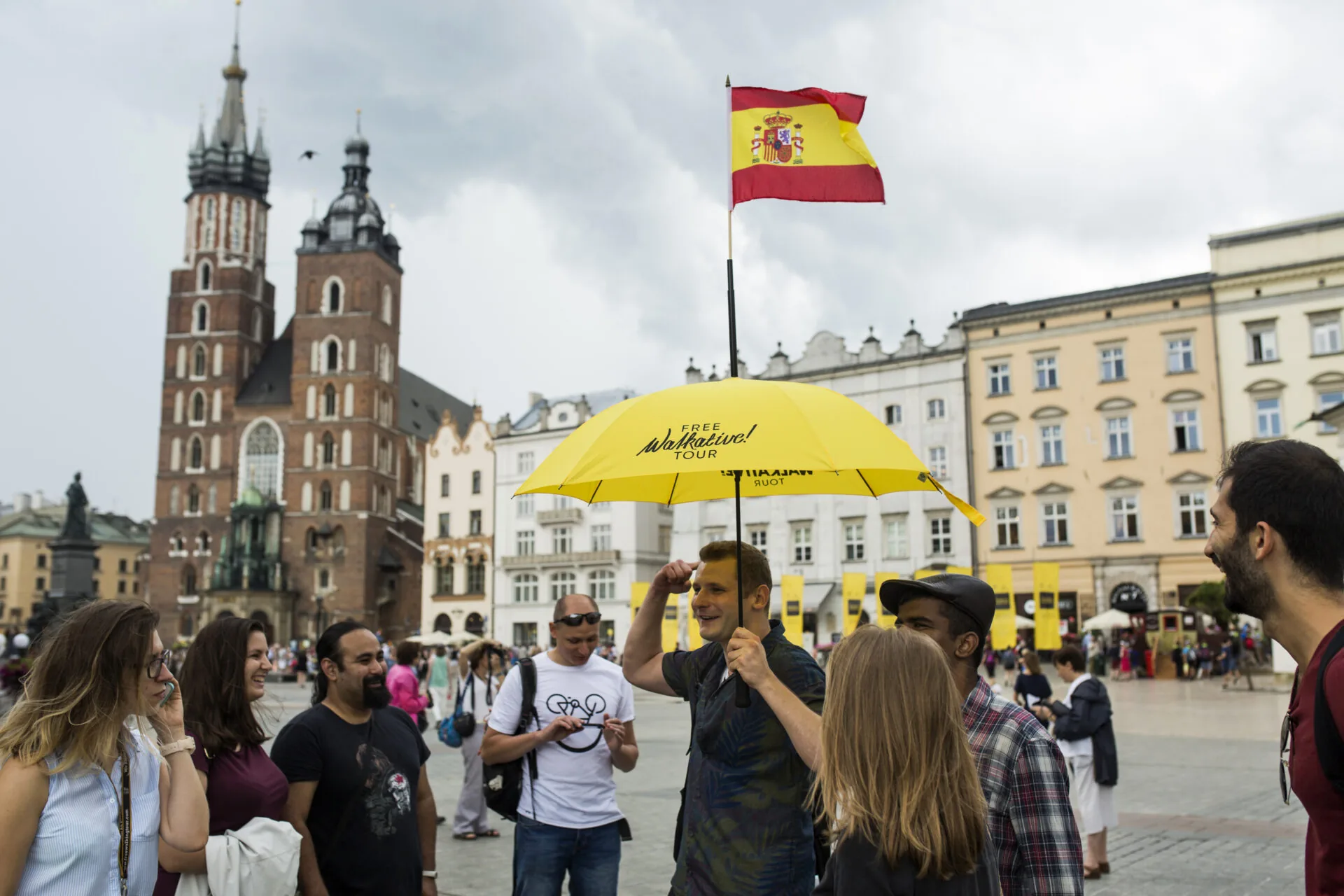 Walkative tour guide with a yellow umbrella and Spanish flag leading a group in Krakow's Main Square.