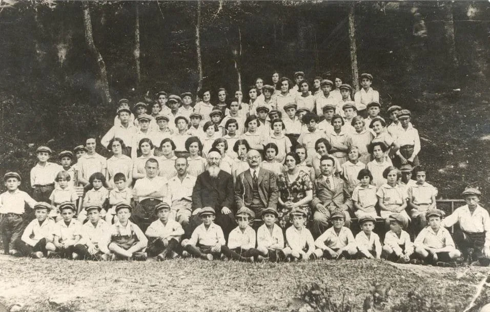 Historical black and white photo of a large group of adults and children posing outdoors in front of trees.