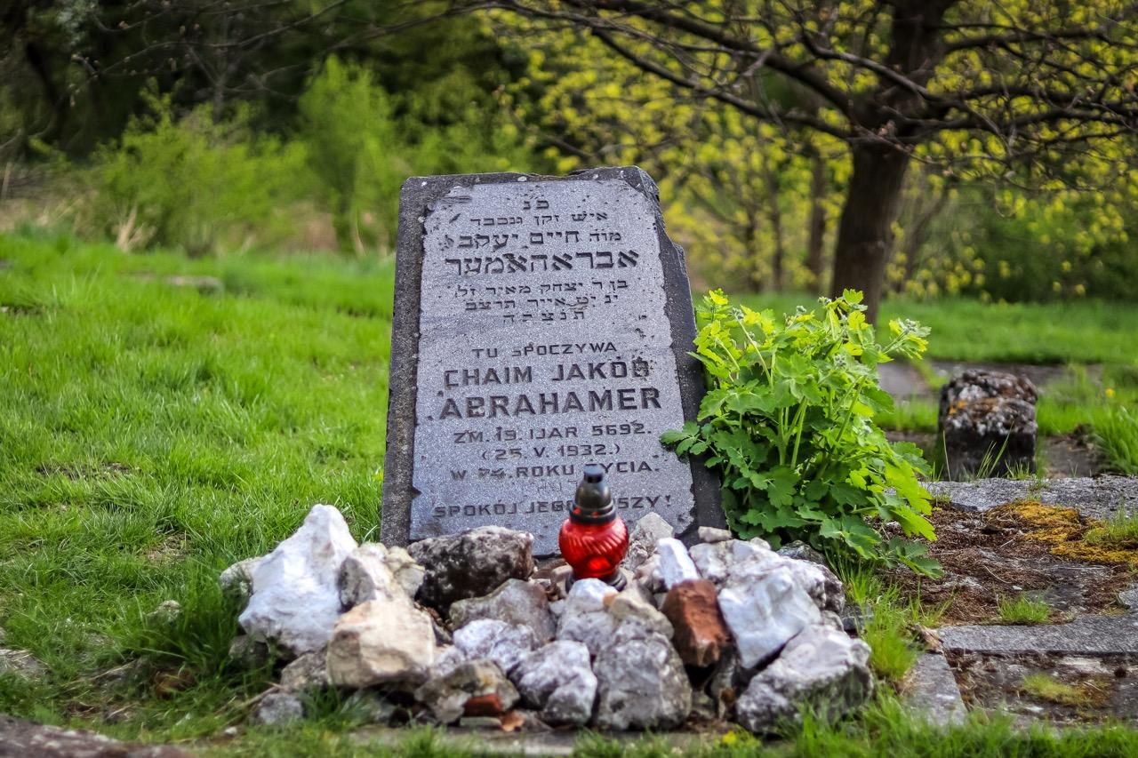 Weathered grey gravestone with Hebrew and Polish text, a red memorial candle, and small stones on grassy ground. Trees in bac