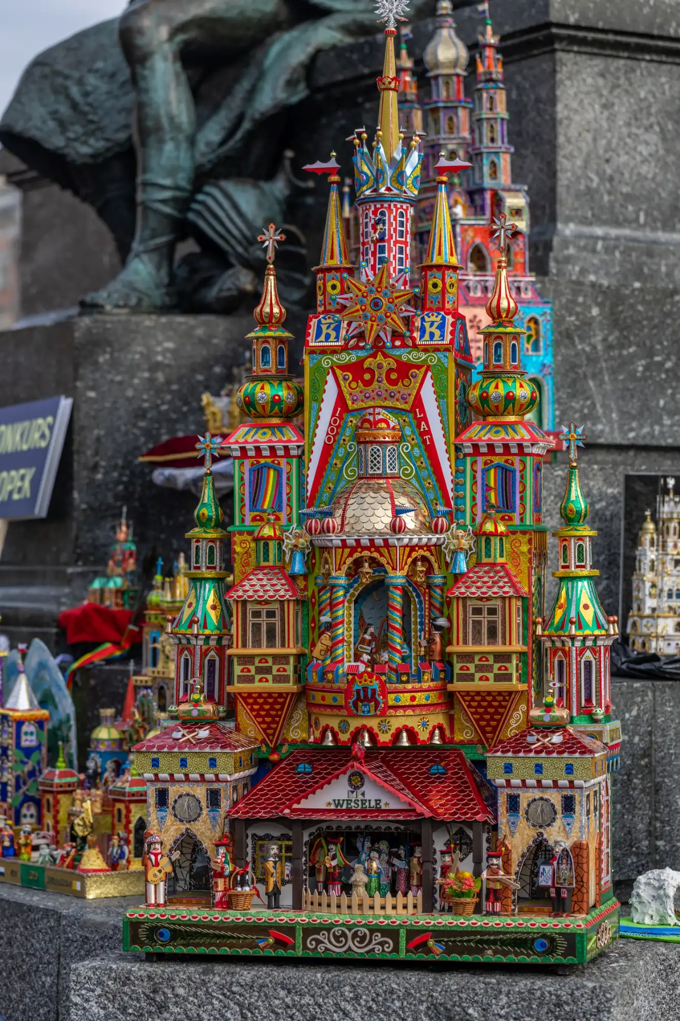 A vibrant, intricate Krakow Szopka (Nativity scene) displayed outdoors, featuring colorful towers, spires, and detailed folk