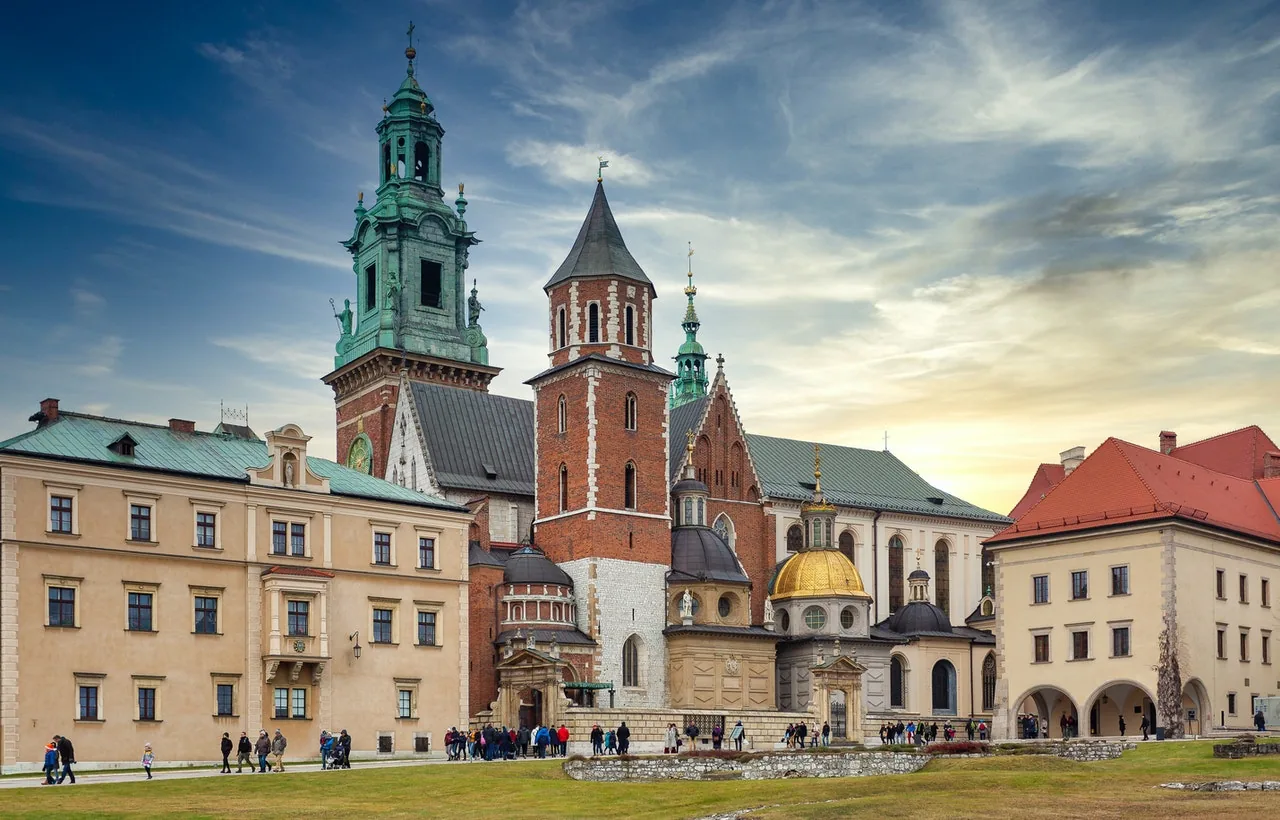 Wawel Cathedral's historic architecture, with green roofs, brick towers, and a golden dome, under a partly cloudy sky. People