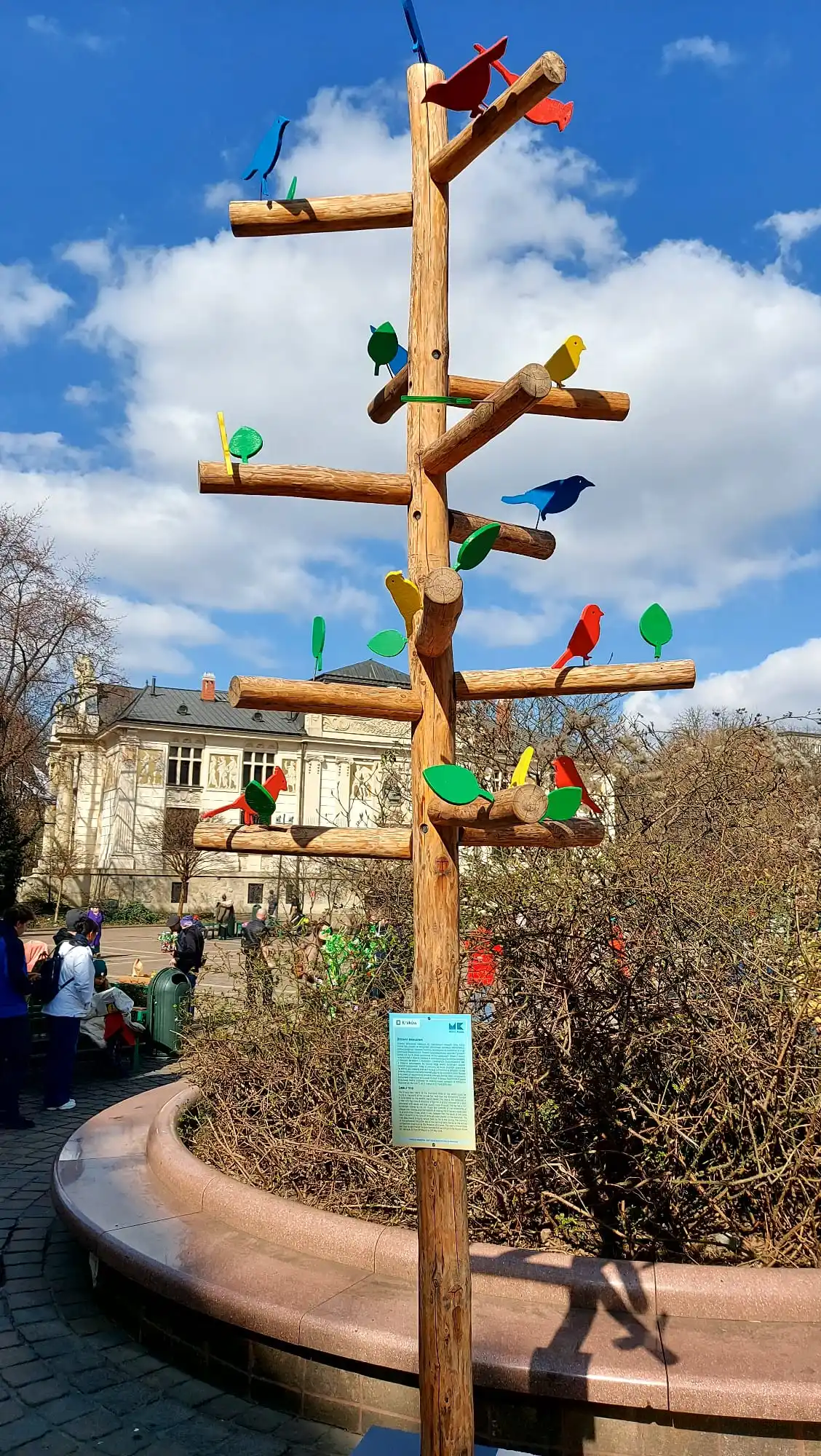 Hohe Holzskulptur in Baumform, mit bunten Vogel- und Blattausschnitten, unter blauem Himmel. Im Hintergrund Menschen und ein