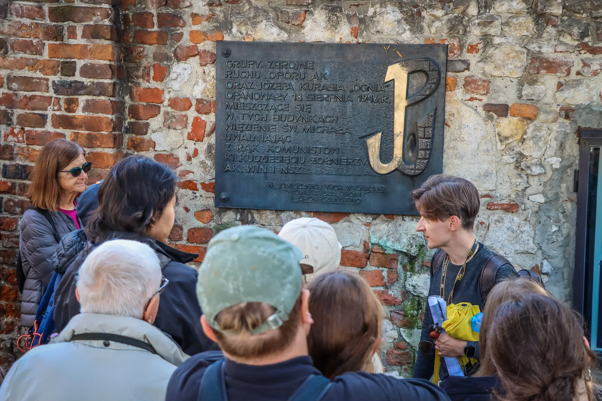 Ein Reiseführer erklärt einer Touristengruppe eine Gedenktafel mit dem Symbol des kämpfenden Polens an einer Ziegelmauer.