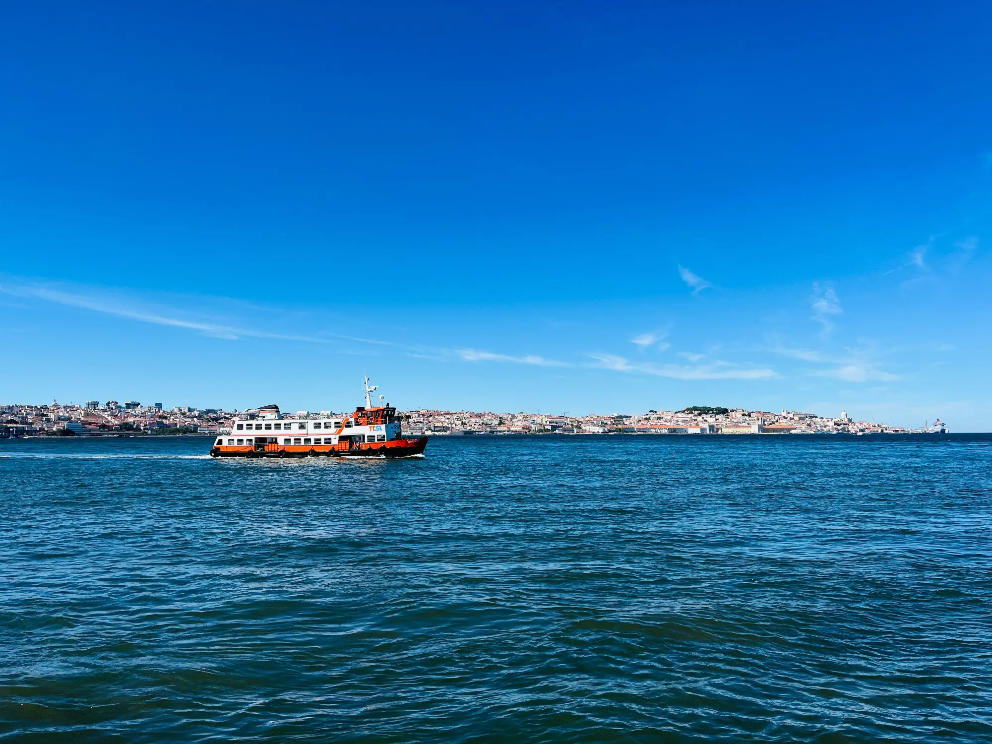 Eine orange-weiße Fähre überquert den Tejo in Lissabon, mit der Stadtsilhouette unter einem klaren blauen Himmel.