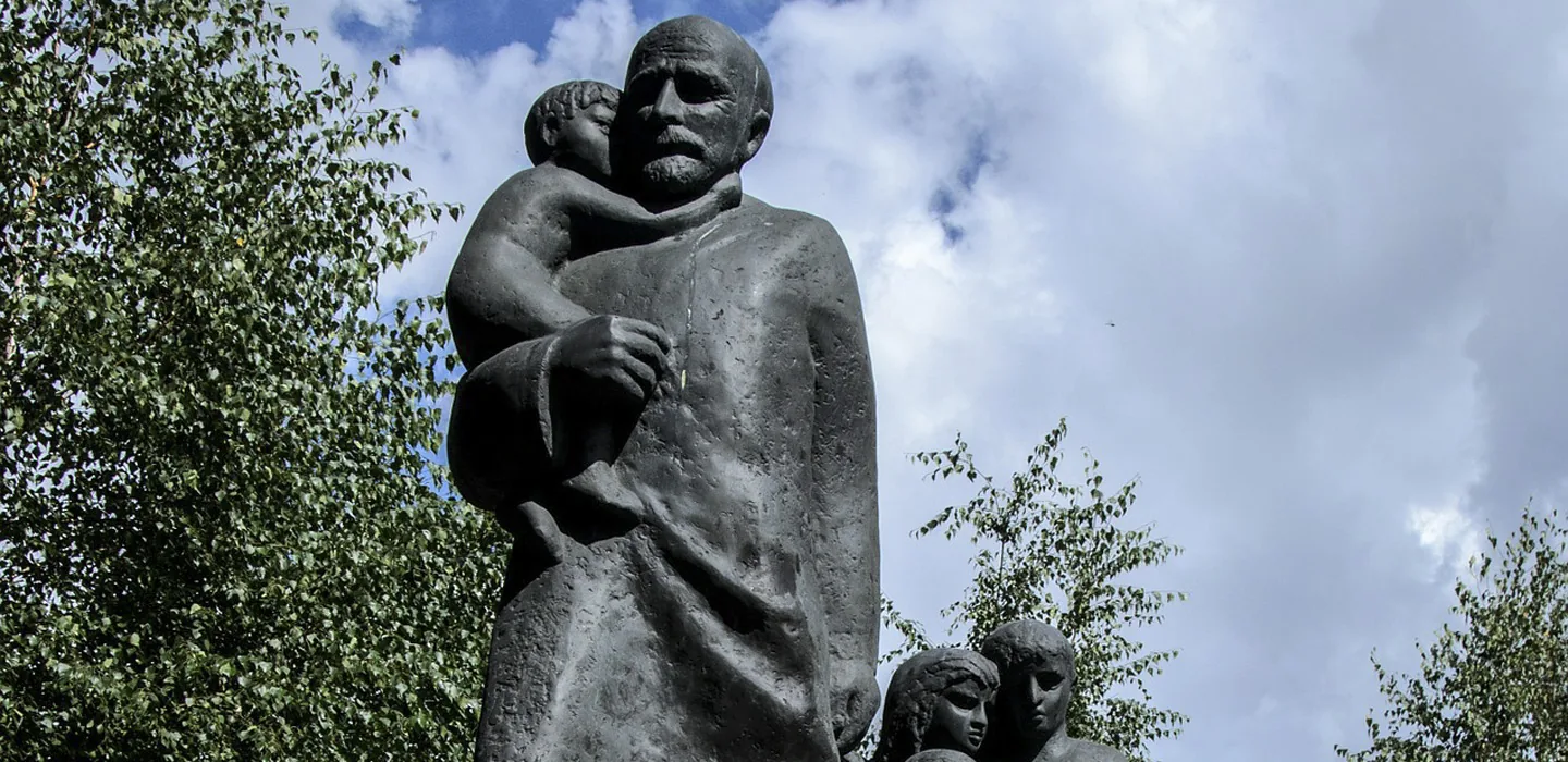 Bronze statue of Janusz Korczak holding a child, with other child figures at the base, against a backdrop of green trees and