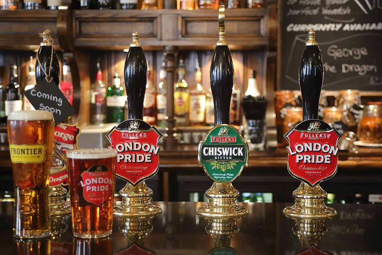 Four beer taps on a dark pub bar counter, two pints of ale, and a 'Coming Soon' sign. Fuller's London Pride and Chiswick Bitt