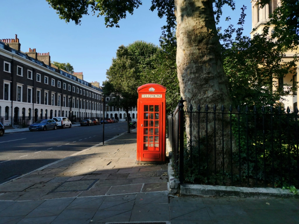 A bright red telephone box stands on a paved sidewalk next to a street with terraced houses and parked cars in London.