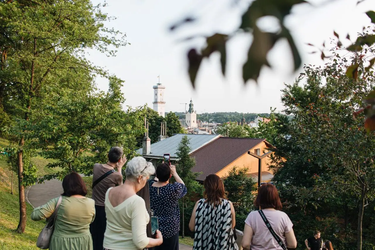 Grupa osób na wzgórzu podziwia panoramę Lwowa z widoczną wieżą Ratusza i kopułą kościoła wśród drzew.
