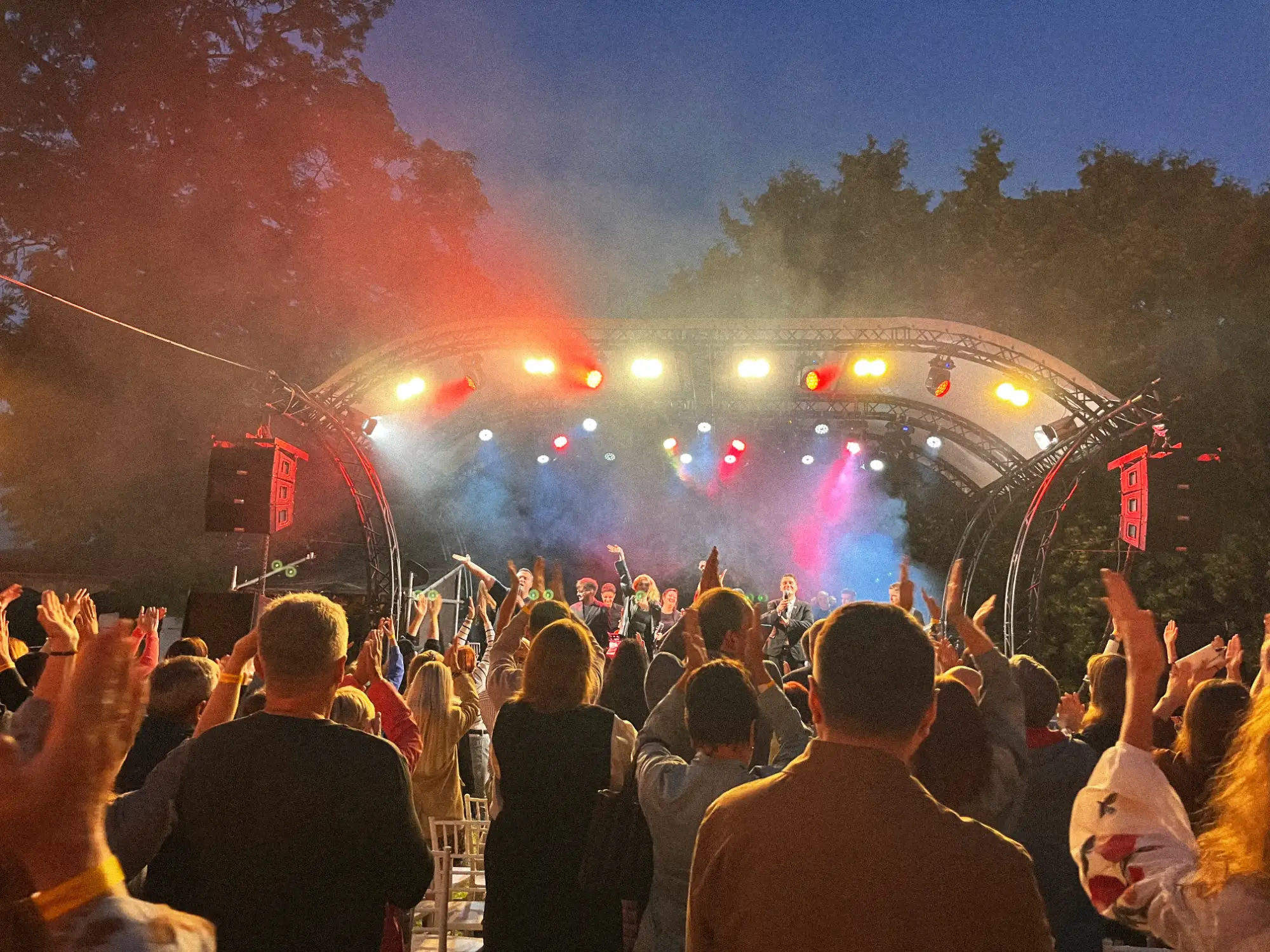 Tłum widziany od tyłu ogląda artystów na jasno oświetlonej scenie plenerowej o zmierzchu.