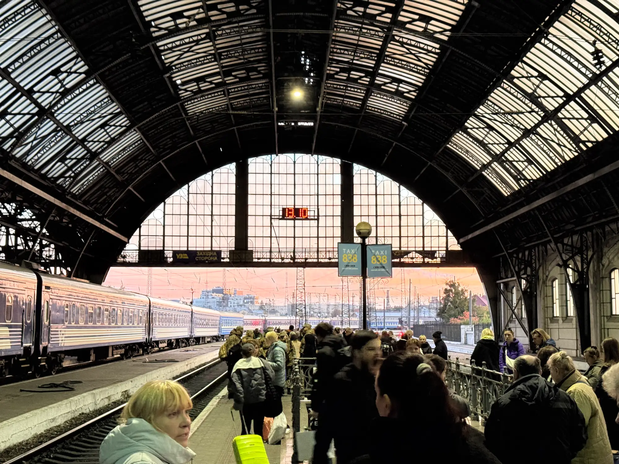 Personas esperan en el andén de una estación de tren junto a un tren azul bajo un gran techo abovedado al atardecer.
