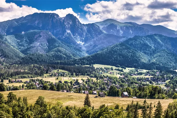 Weitläufiges Gebirge mit dichten grünen Wäldern an den Hängen, das in ein Tal mit verstreuten Häusern und Feldern unter bewöl
