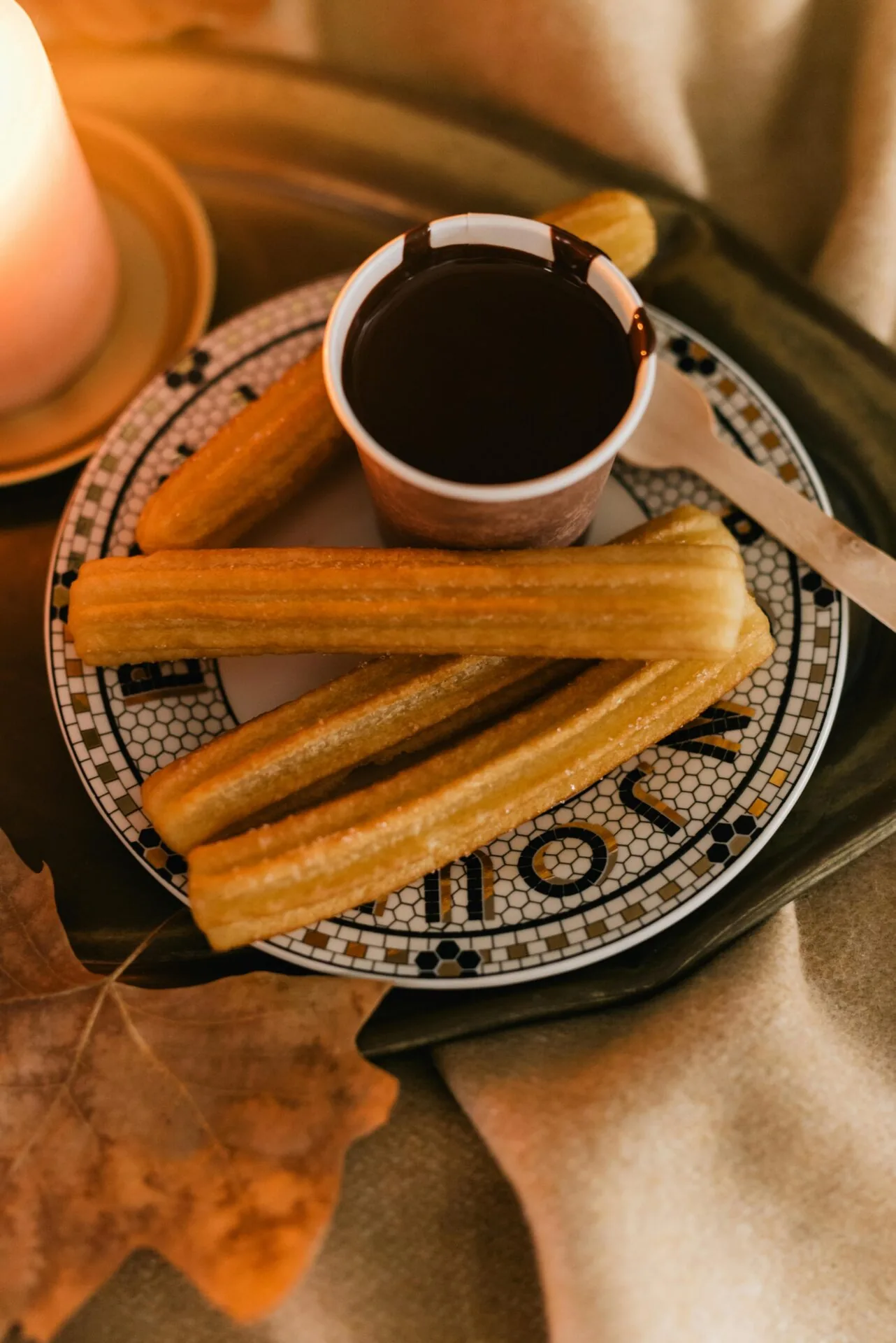 Plate with several sugar-dusted churros, a cup of thick dark chocolate, a wooden spoon, a lit candle, and an autumn leaf.
