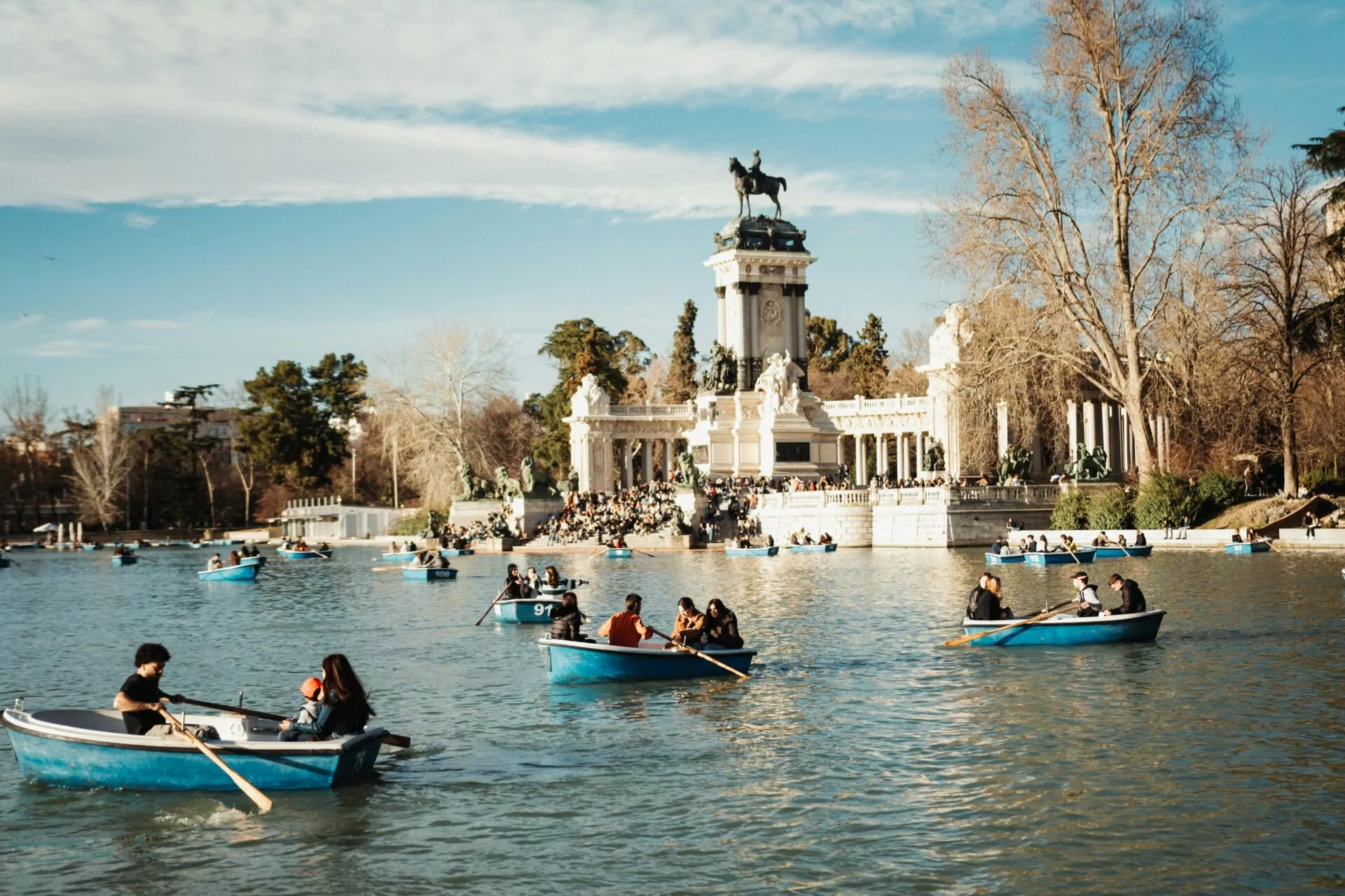People in blue rowboats on a lake with the Alfonso XII monument and trees in Madrid's Retiro Park.