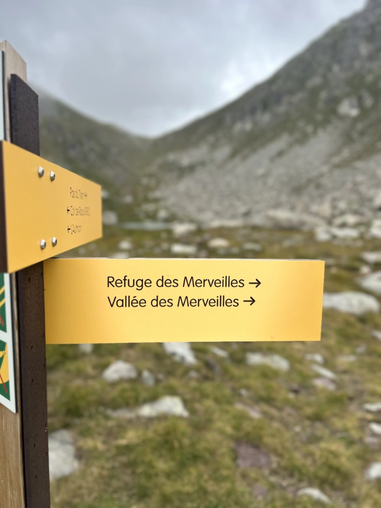 Yellow hiking signpost in a rocky mountain landscape under a cloudy sky, pointing to 'Refuge des Merveilles' and 'Vallée des