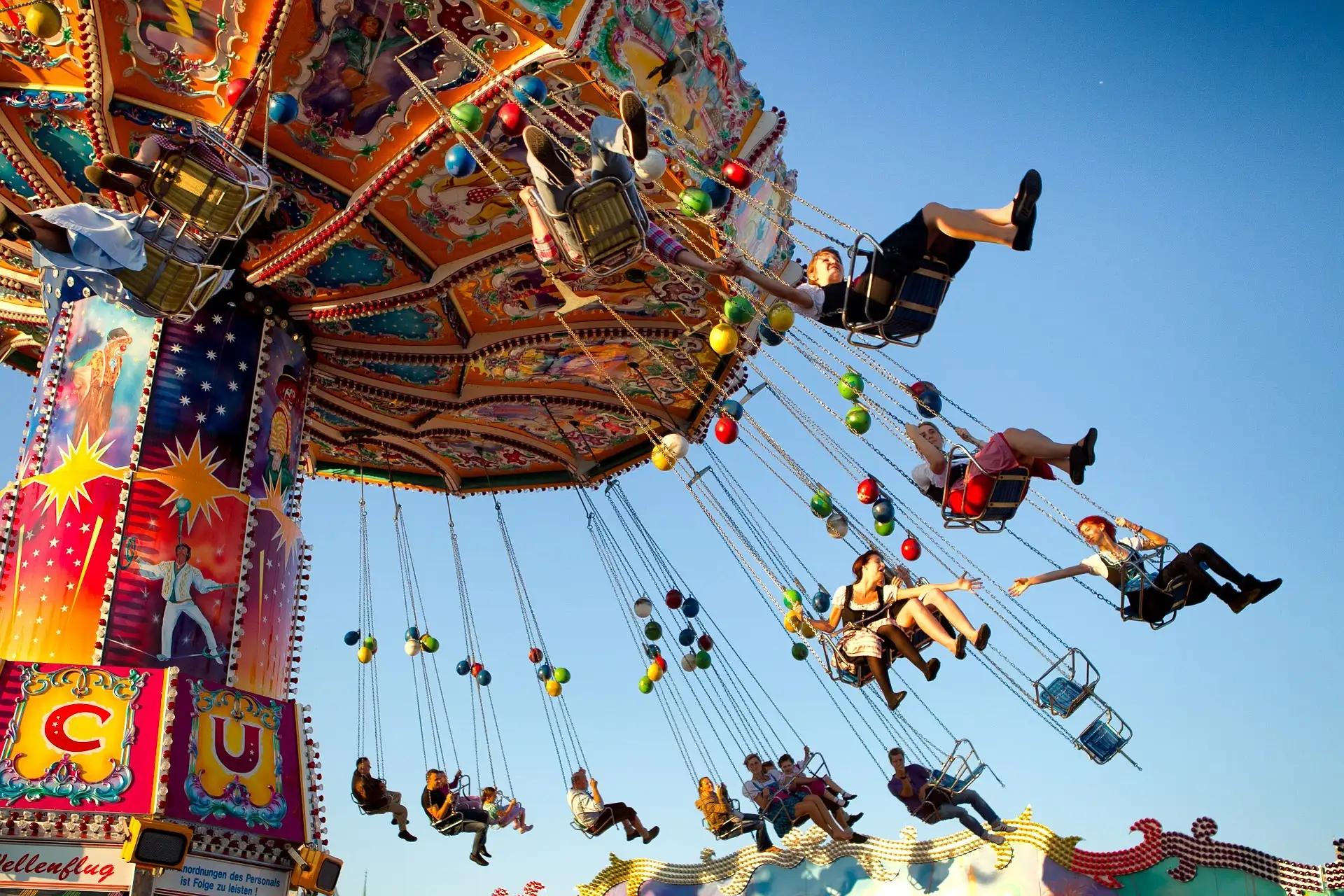 Buntes Kettenkarussell mit mehreren Personen, die vor blauem Himmel in der Luft schweben. Der dekorierte Sockel des Fahrgesch