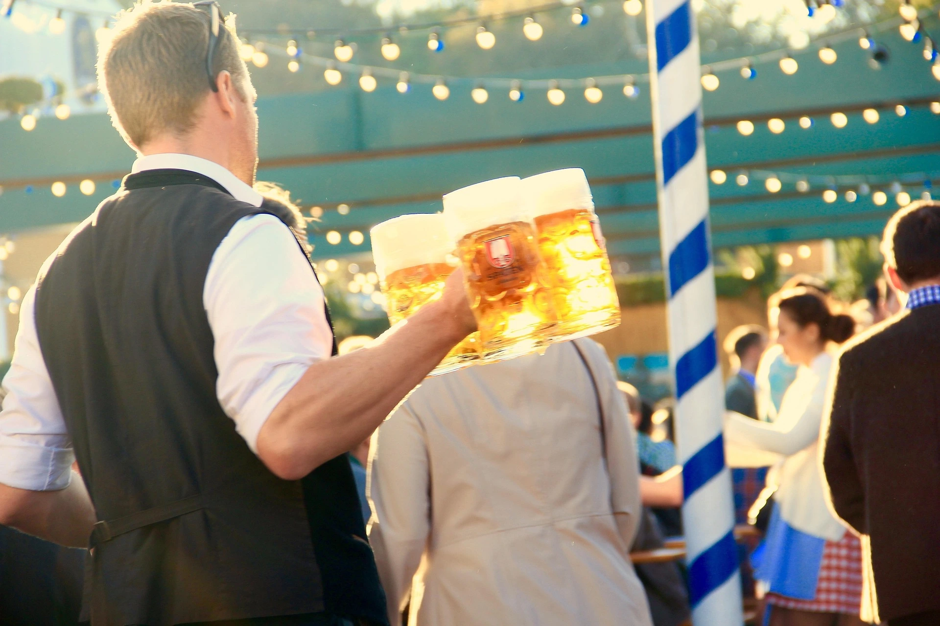 Una persona lleva tres grandes jarras de cerveza espumosa. Al fondo, personas borrosas, luces y un poste a rayas azules y bla