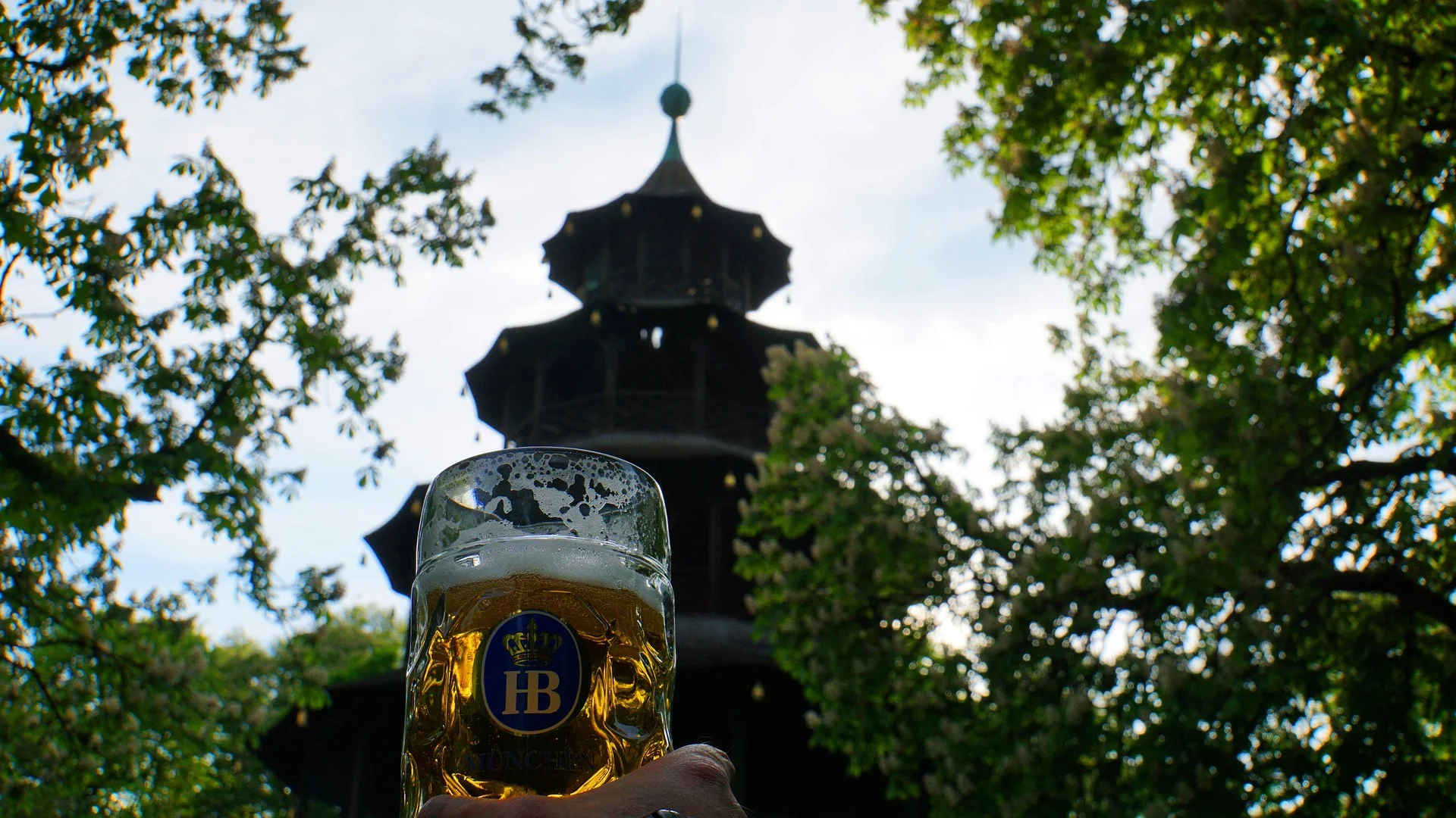 Eine Hand hält einen vollen Bierkrug mit HB-Logo im Vordergrund, dahinter der unscharfe Chinesische Turm und grüne Bäume.