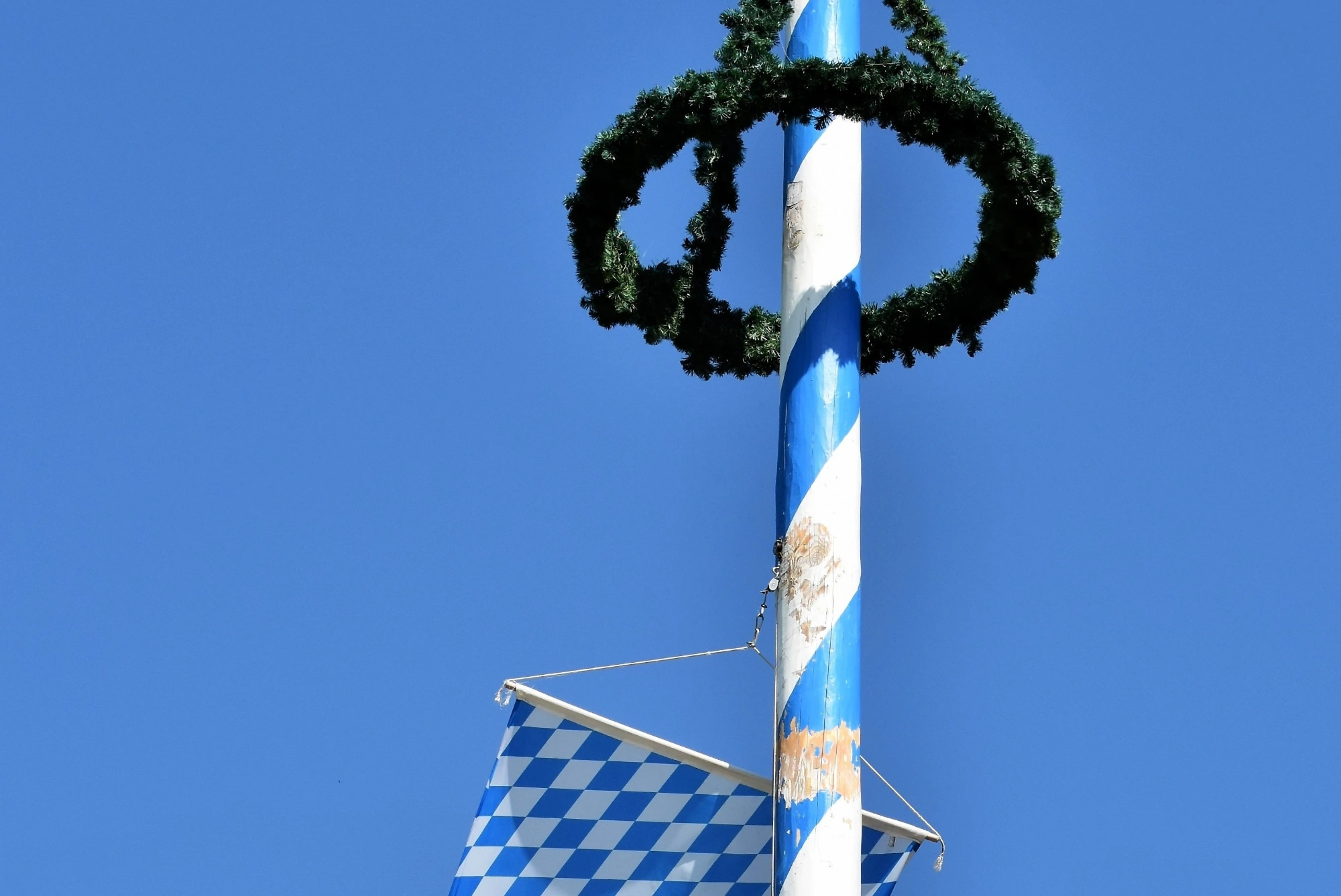 Blue and white maypole with a green wreath and Bavarian flag against a clear sky