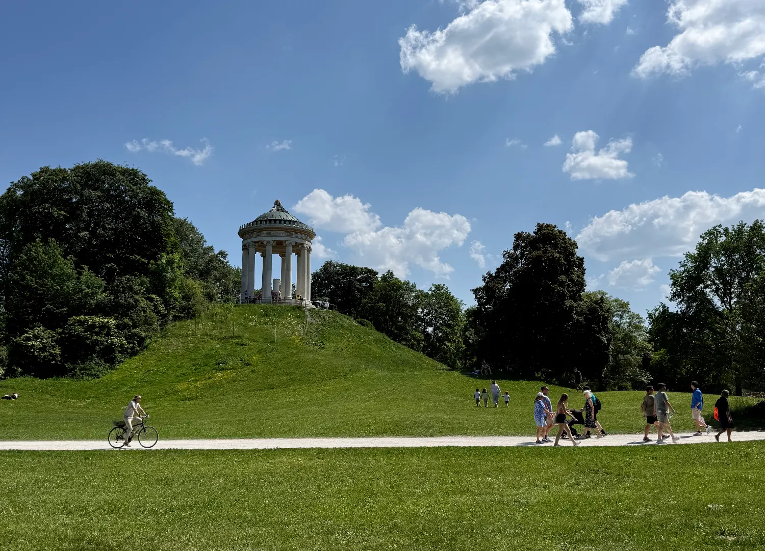 Der Monopteros-Tempel auf einem grünen Hügel im Englischen Garten, mit Menschen, die auf dem Weg spazieren gehen.