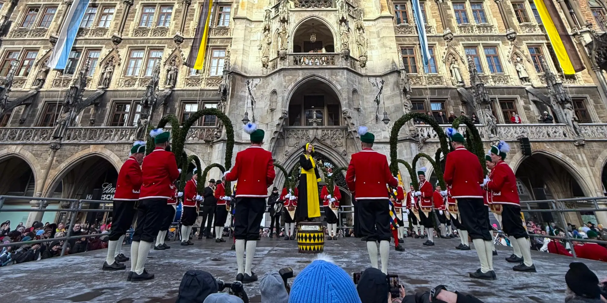 Men in traditional costumes perform the Schaefflertanz in front of Munich's Neues Rathaus, with a woman in a yellow dress.