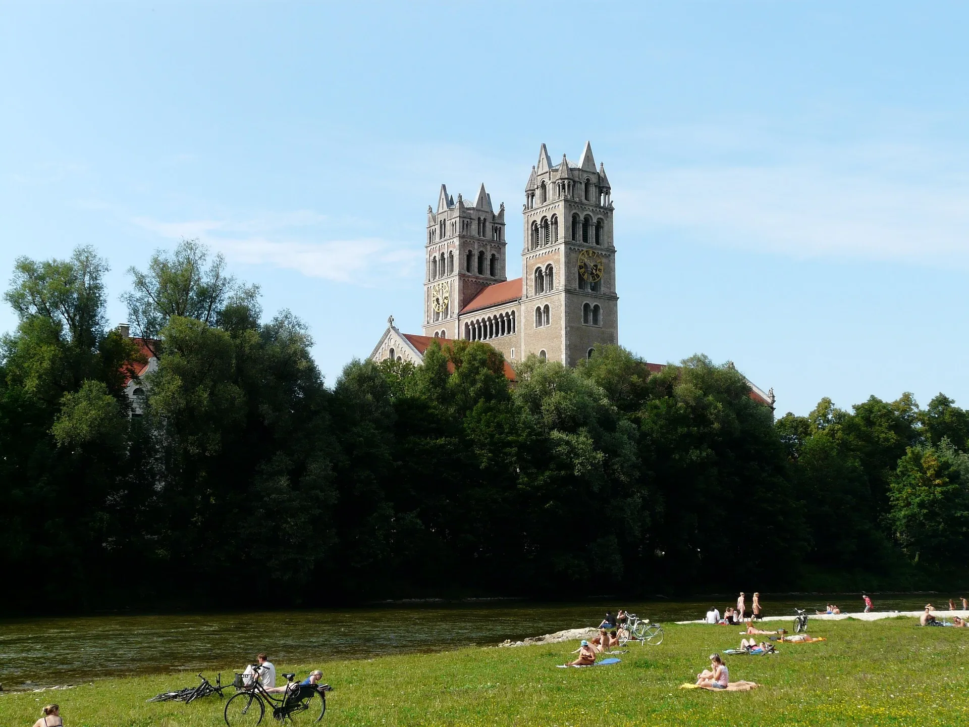 St. Maximilianskirche mit zwei Türmen und rotem Dach, über Bäumen vom Grasufer der Isar aus gesehen, wo Menschen entspannen.