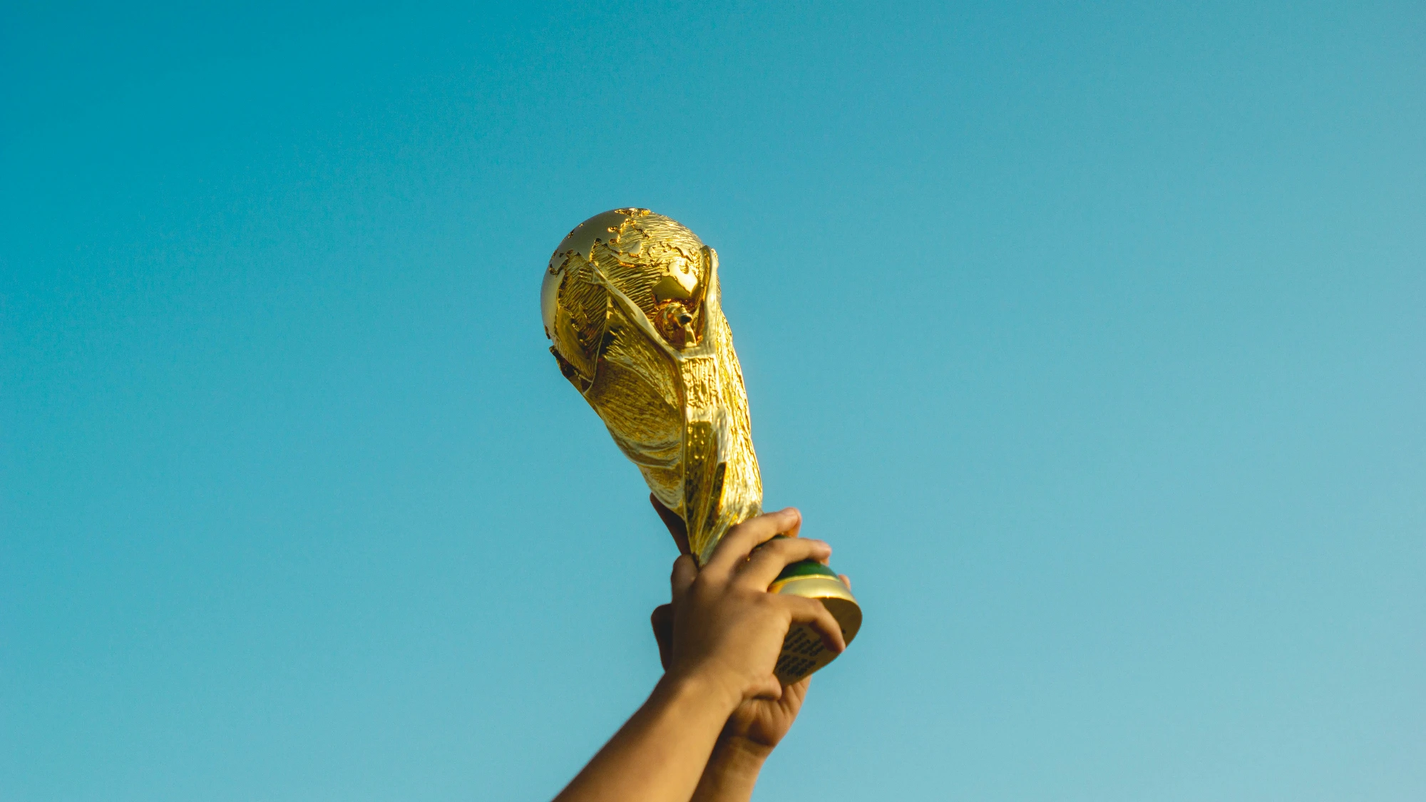 Hands holding the golden FIFA World Cup trophy high against a clear blue sky.