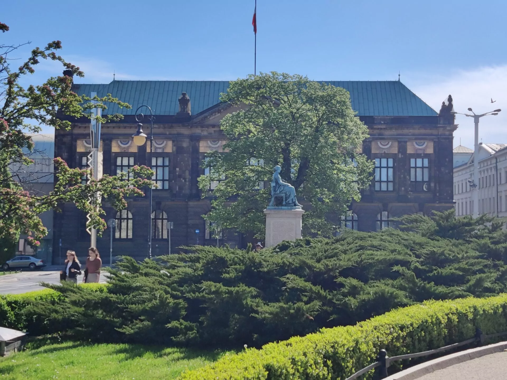 Blick auf das Nationalmuseum in Posen mit einer sitzenden Statue im Vordergrund, umgeben von grünen Büschen und Bäumen.