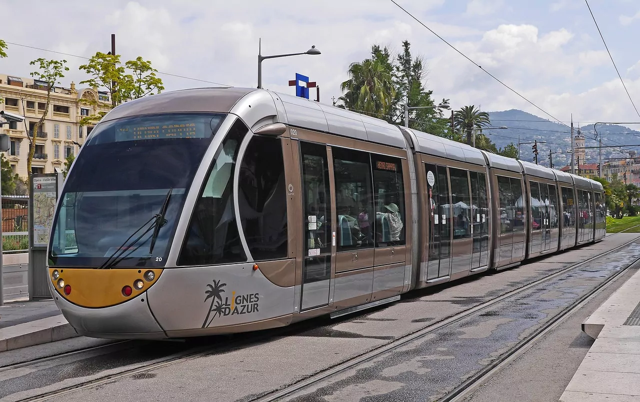 Eine moderne silber-braune Straßenbahn mit "Lignes d'Azur"-Logo auf Gleisen in einer Stadtstraße. Gebäude und Palmen sind im