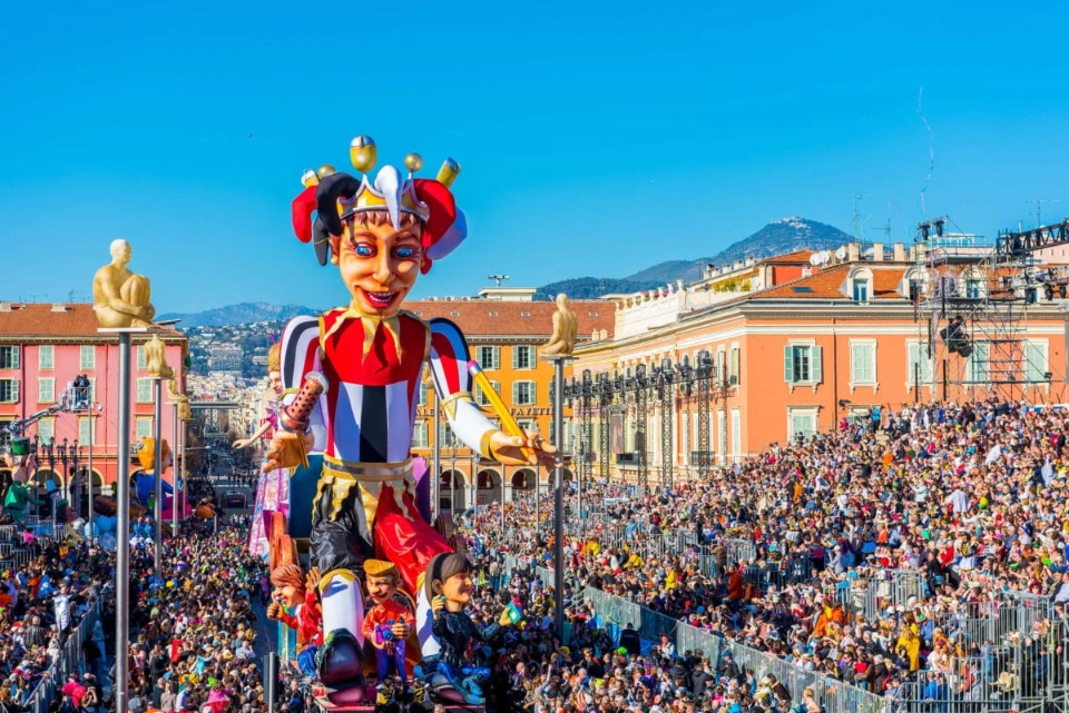 Large jester parade float with a huge crowd of spectators in a sunny city square. Buildings and mountains are visible.