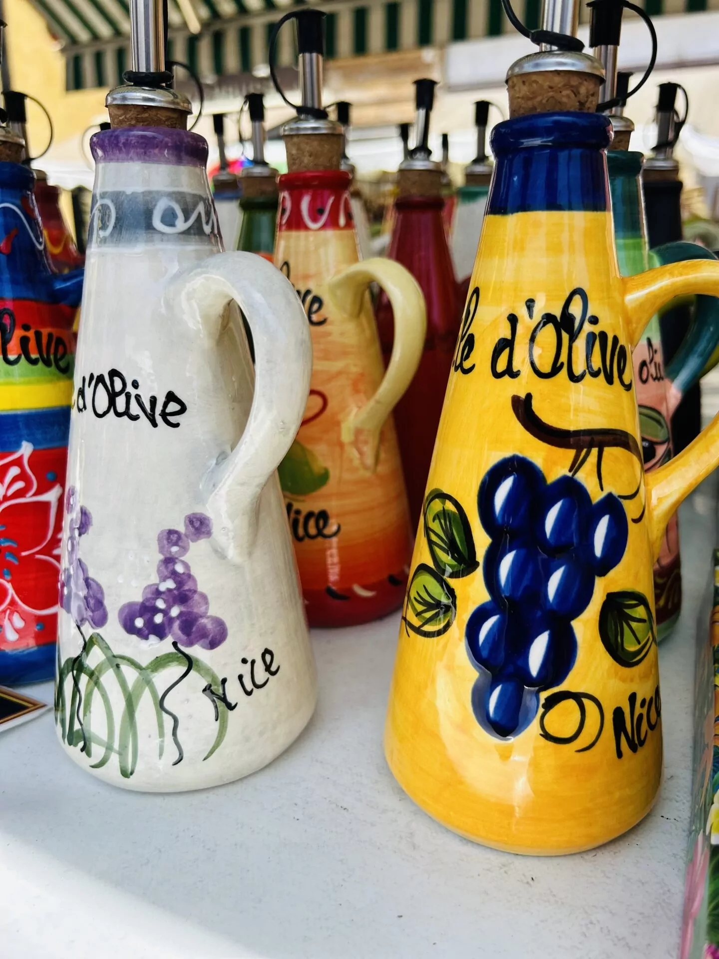 Zbliżenie na kolorowe ceramiczne butelki na oliwę z dozownikami, ozdobione winogronami i kwiatami, na białej powierzchni.
