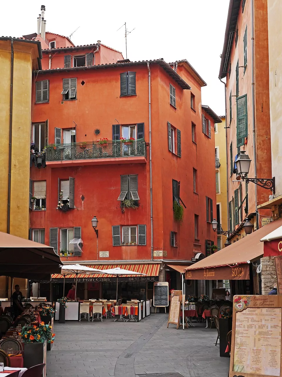 Enge Gasse in der Altstadt von Nizza mit terrakottafarbenen Gebäuden, geöffneten Fensterläden und Restaurants mit Außensitzpl