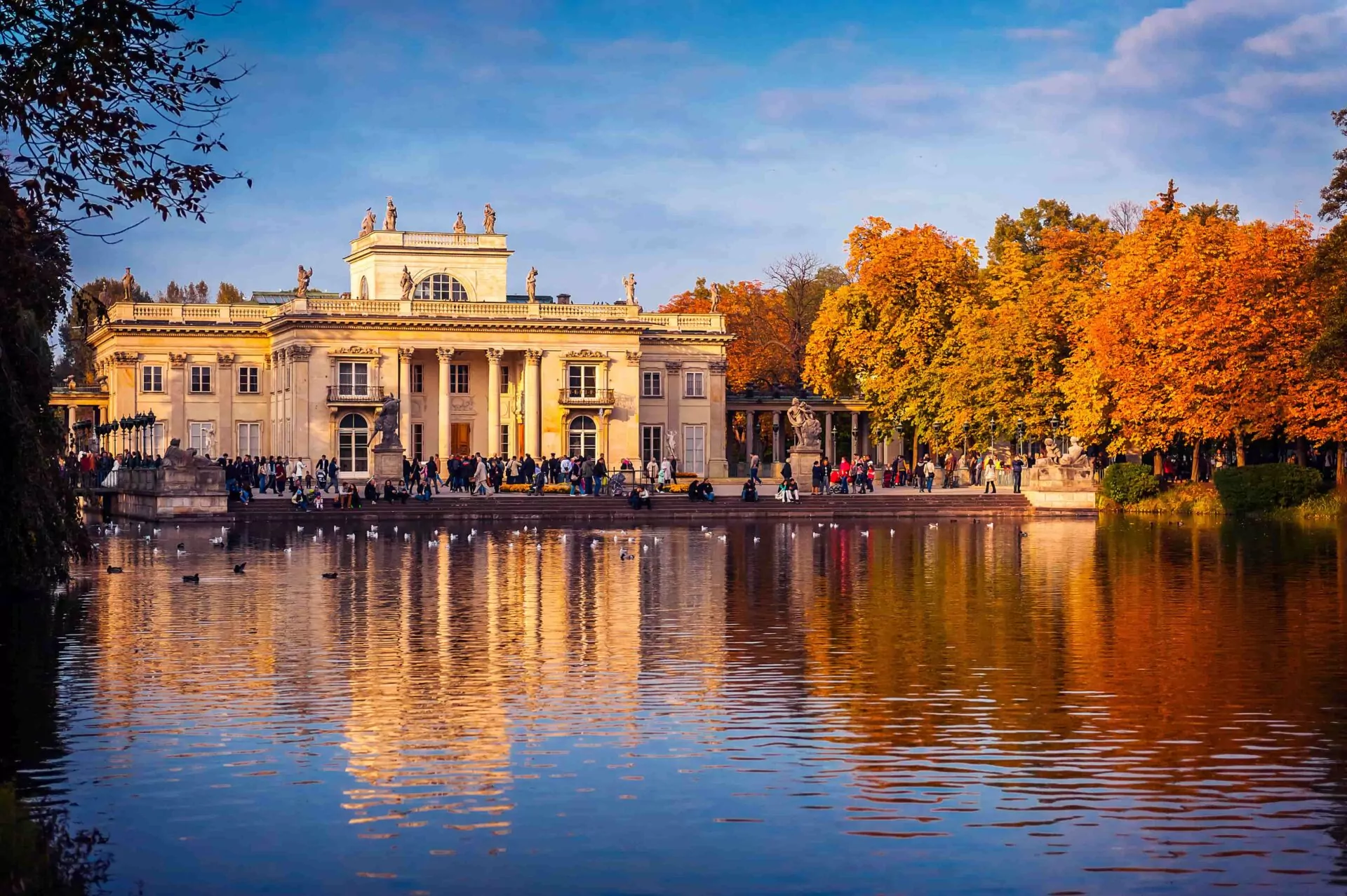 The Palace on the Isle in Łazienki Park, Warsaw, viewed across a lake with ducks. People gather on the shore among autumn tre