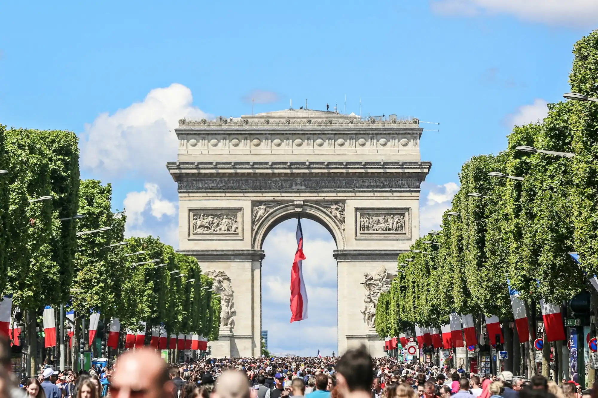 View of the Arc de Triomphe from a crowded Champs-Élysées, lined with French flags for a national celebration or parade.