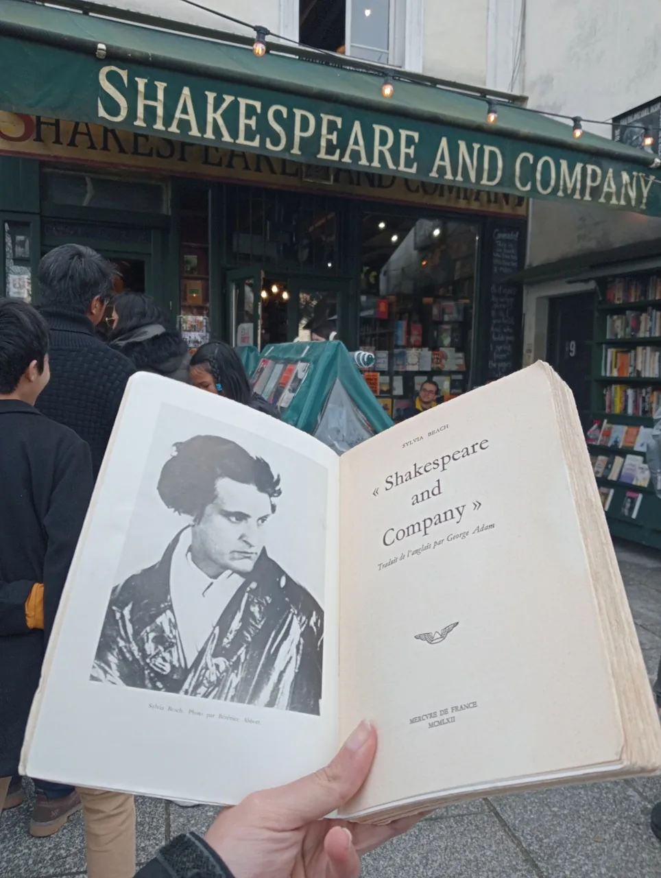 Hand holding open book with Sylvia Beach photo in front of Shakespeare and Company bookstore in Paris, showing other visitors
